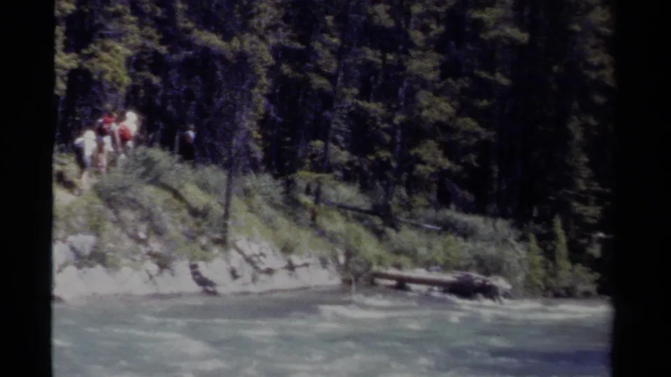 A distant group of hikers appears on a forested British Columbia riverbank in soft, grainy 8mm footage.