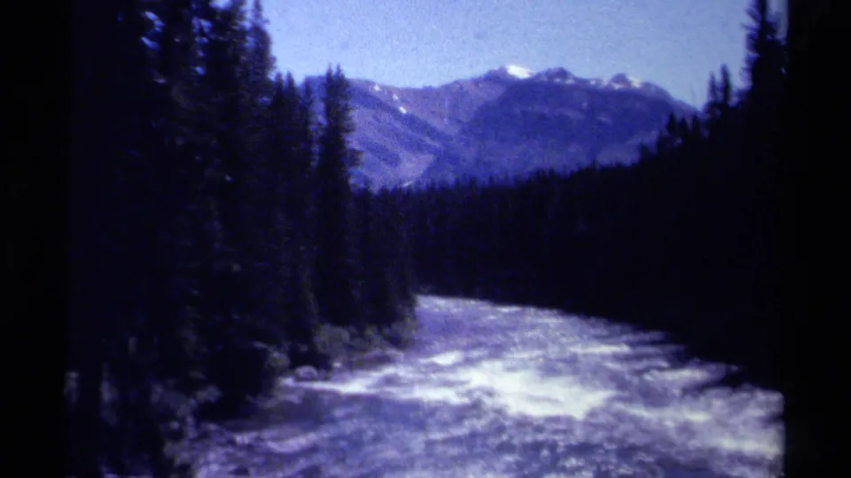 A wide 8mm view looks down a fast British Columbia river toward forested slopes and distant snow-touched mountains.