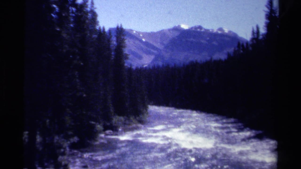 British Columbia 1979 8mm frame of a bright river running through dark evergreen forest toward distant mountain peaks.