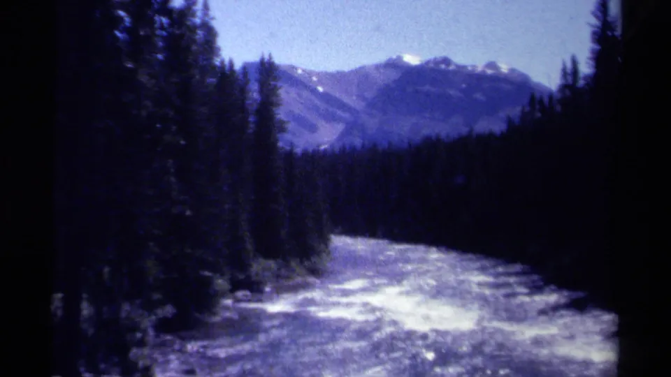 British Columbia 1979 8mm frame of a bright river running through dark evergreen forest toward distant mountain peaks.