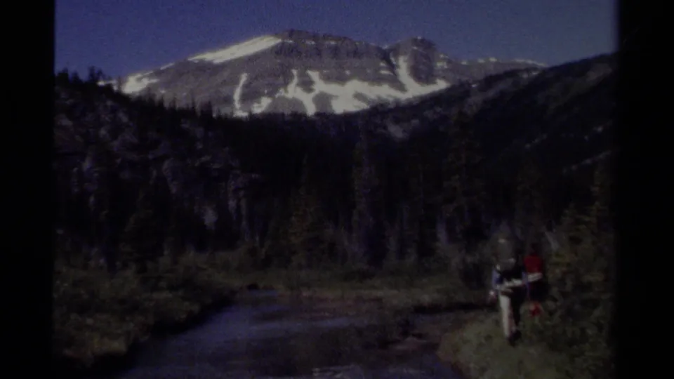 Two distant backpackers walk along a riverbank beneath snow-dusted mountains in British Columbia, Canada, in 1979 8mm footage.