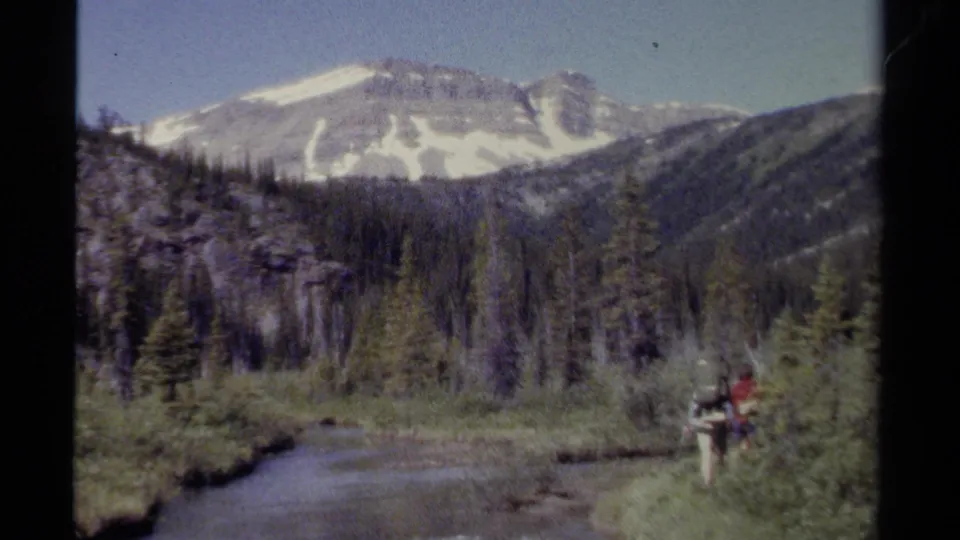Distant hikers with backpacks pause near a river in a forested British Columbia mountain landscape, filmed on 8mm in 1979.