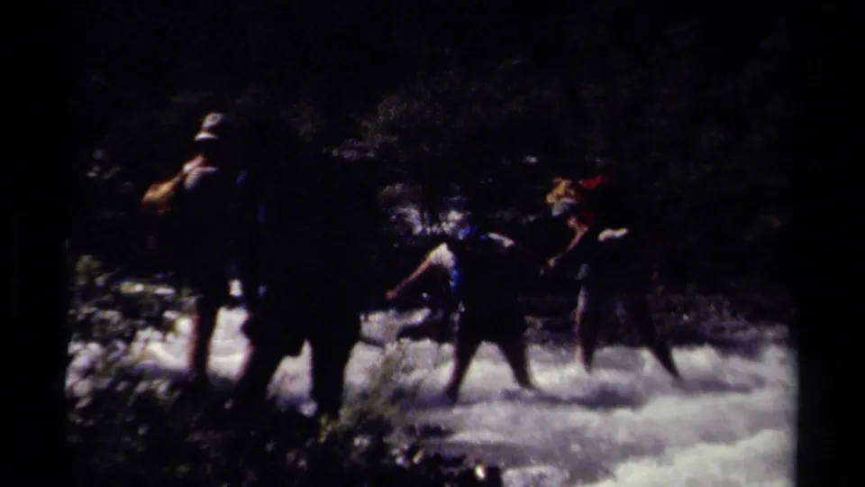 Backpacked hikers in British Columbia appear to cross a fast mountain stream, partly silhouetted by bright water and forest shade.