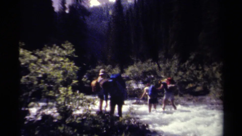 1979 8mm frame of several backpacked hikers wading across a fast stream in a forested British Columbia mountain setting.