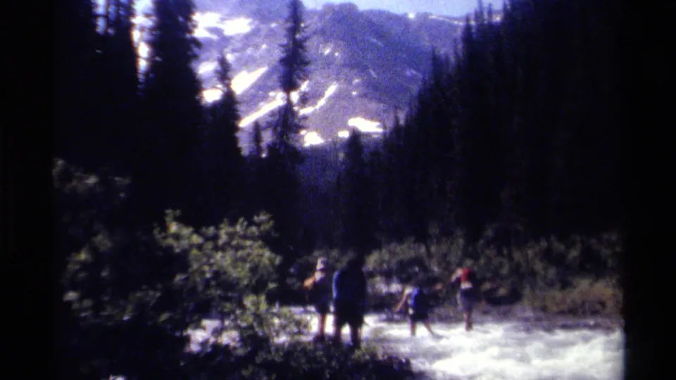 Backpacked hikers wade through whitewater as forest and distant snowy mountain slopes frame the British Columbia wilderness scene.