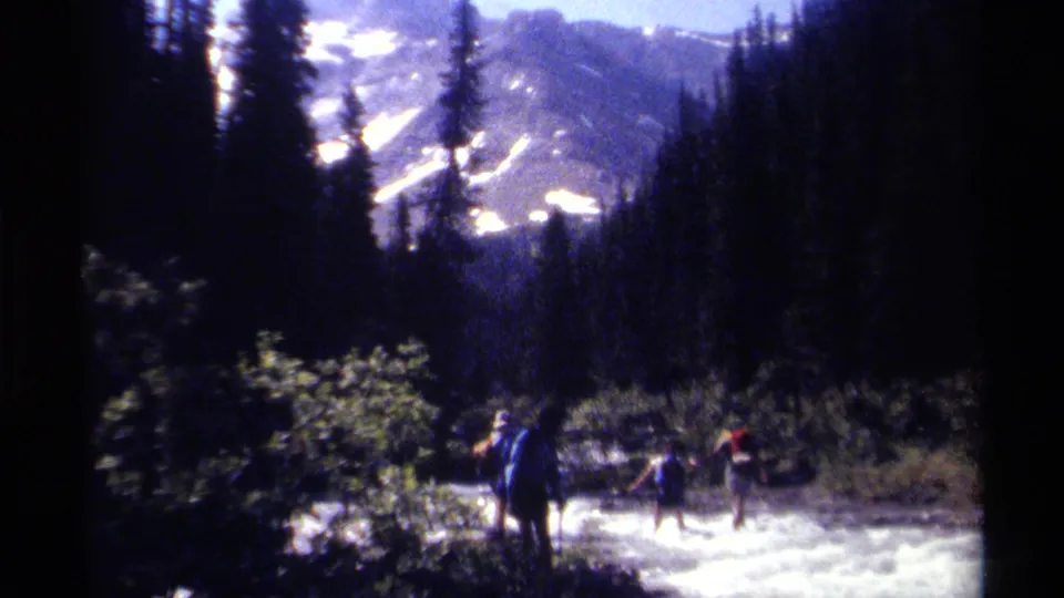 1979 British Columbia 8mm frame showing distant hikers crossing a rushing stream in a forested mountain valley.