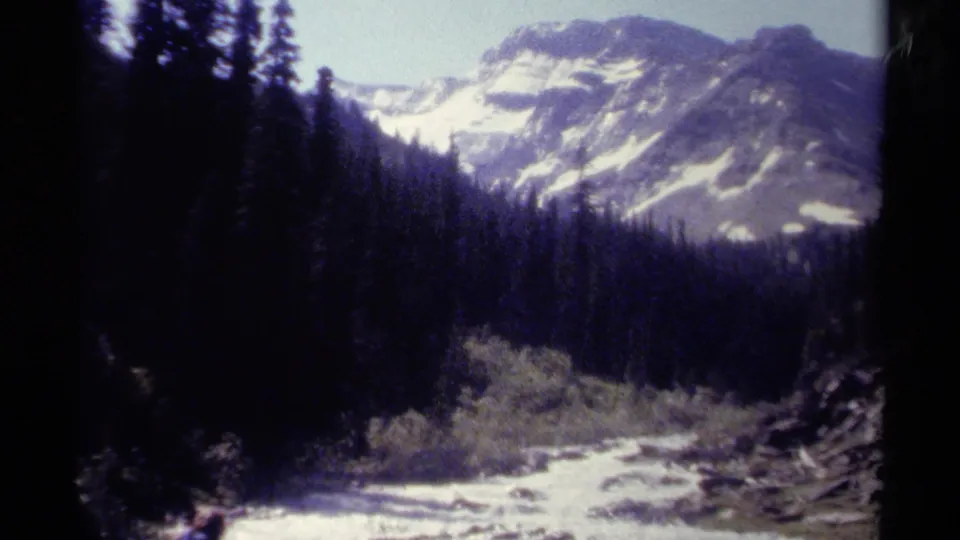 1979 British Columbia 8mm frame looking across a bright stream toward dense evergreens and snowy mountain slopes.