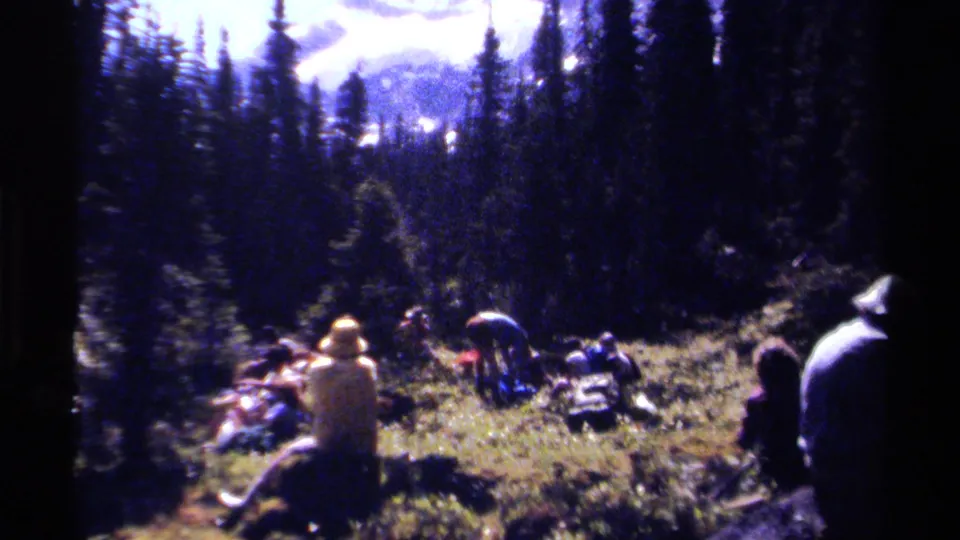 1979 British Columbia camping scene showing people seated and standing in a meadow among conifers, with outdoor gear and mountains in the background.