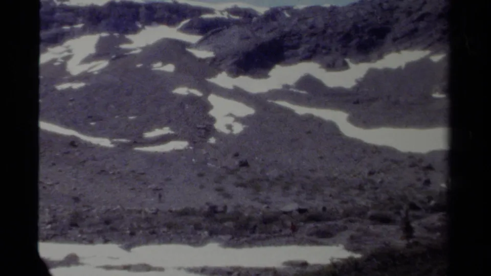 British Columbia mountain terrain on 8mm film, with snow patches and a tiny red-clad figure visible against the rocky slope.
