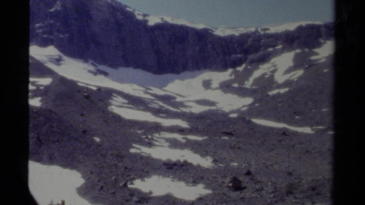 1979 British Columbia mountain view with a jagged dark ridge, scattered snowfields, and visible 8mm scan texture.