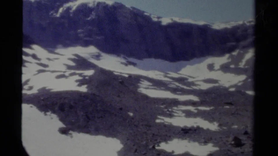 1979 British Columbia home-movie frame of isolated rocky peaks and snowfields under a clear pale sky.