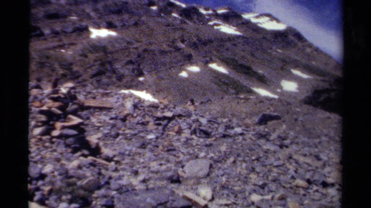 Rocky mountain slope with snow patches and a small stone cairn.