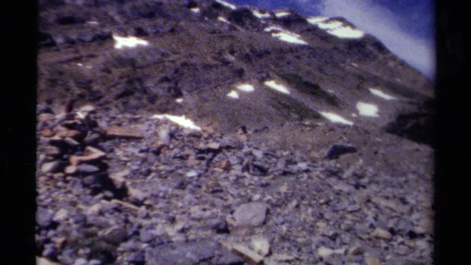 1979 British Columbia 8mm frame showing a rocky alpine slope with snow patches and a small cairn in the foreground.