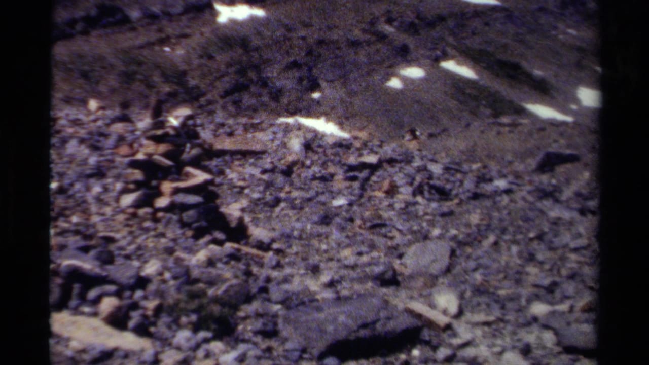 1979 British Columbia home-movie frame with a stacked stone cairn on gravel below snow-streaked mountain terrain.
