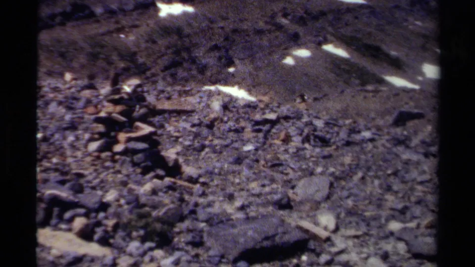 1979 British Columbia home-movie frame with a stacked stone cairn on gravel below snow-streaked mountain terrain.