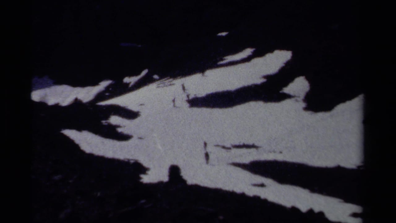 Grainy 8mm frame from British Columbia showing a shadowed alpine slope with irregular snow patches and distant hikers.