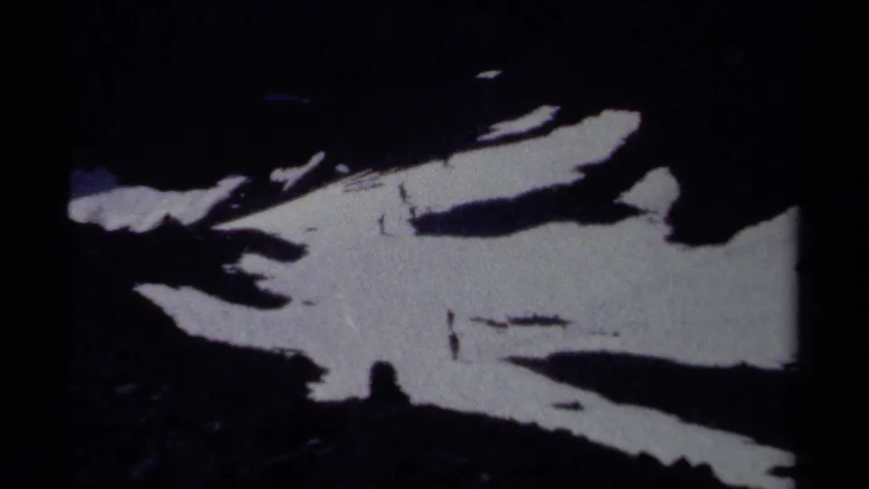 Grainy 8mm frame from British Columbia showing a shadowed alpine slope with irregular snow patches and distant hikers.