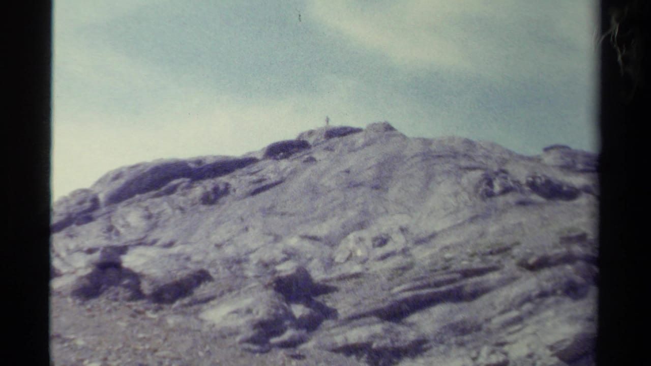 Faded color 8mm frame of a rocky British Columbia mountain ridge with a tiny summit figure under a pale sky.