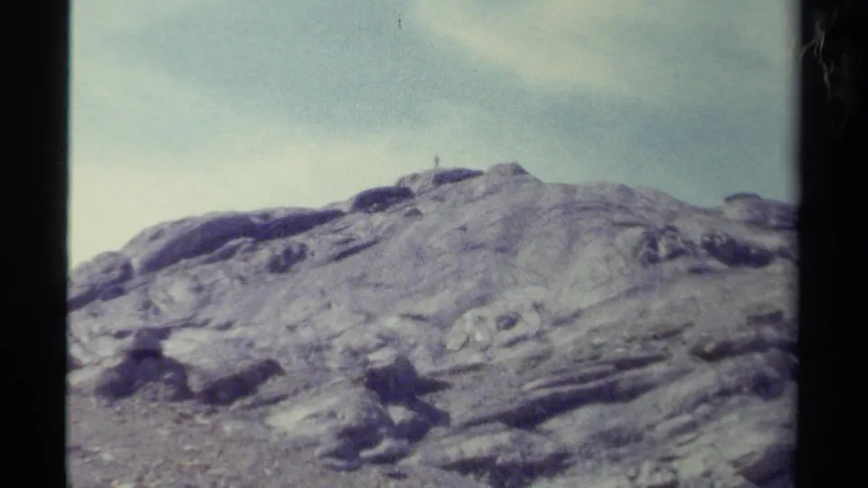 Faded color 8mm frame of a rocky British Columbia mountain ridge with a tiny summit figure under a pale sky.