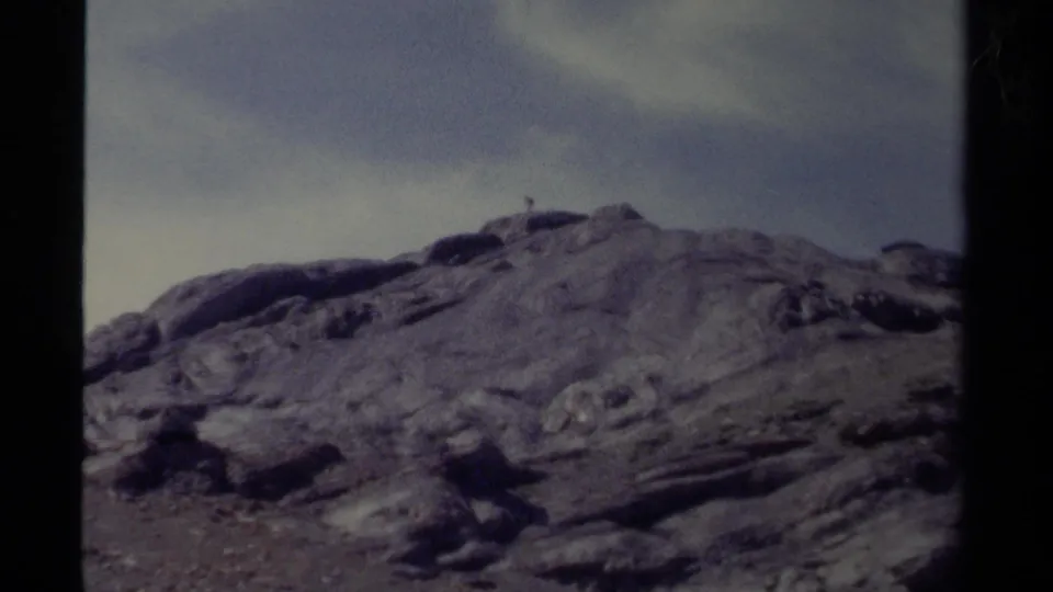 Vintage 8mm view of a British Columbia alpine ridge with rough rock surfaces, pale sky, and faint distant figure detail.