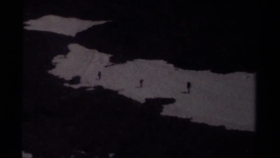 Dark, grainy 8mm frame from British Columbia showing hikers as small silhouettes on a snowfield in alpine terrain.