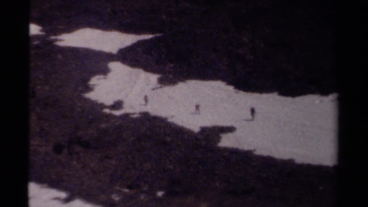 Distant hikers cross a snow patch on rocky mountain terrain.