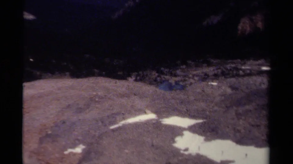Grainy 8mm frame shows distant hikers near snow patches on uneven alpine terrain in British Columbia.