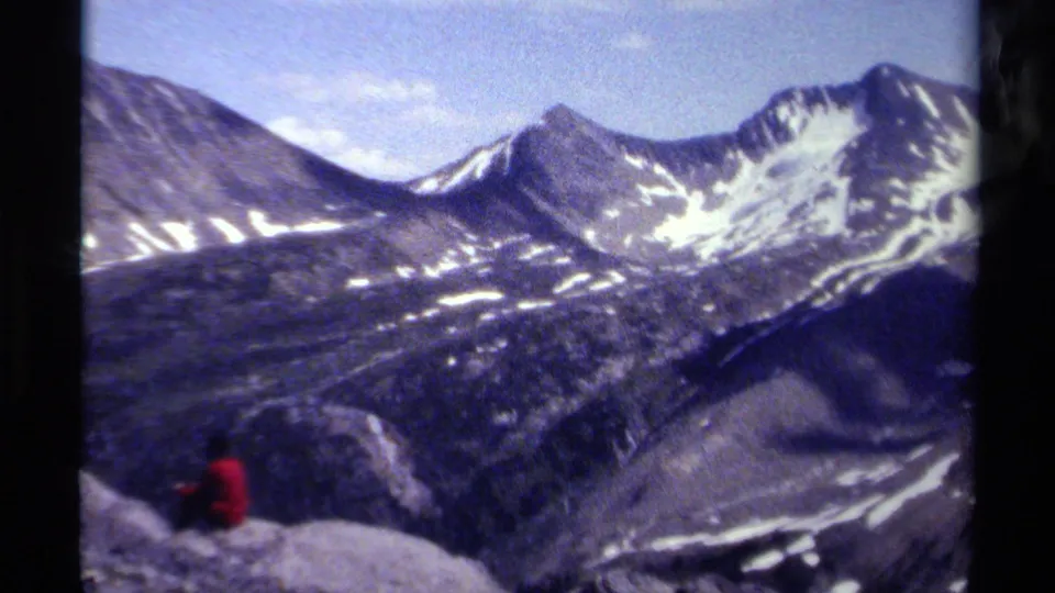 1979 8mm British Columbia alpine scene with snow-streaked peaks and a small seated figure in a red jacket at lower left.