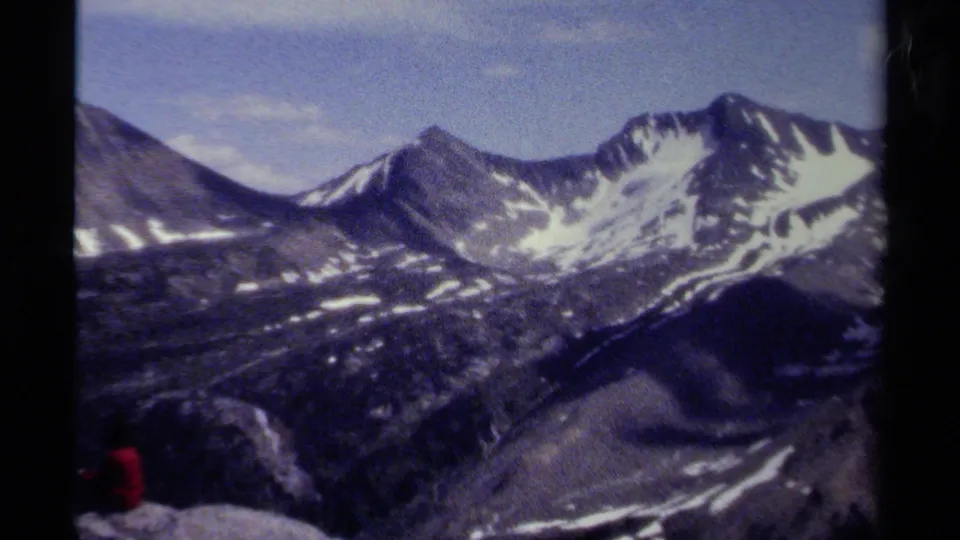 Wide 1979 8mm British Columbia mountain panorama with snow-striped slopes, pale sky, and a small seated figure in red near the lower left.