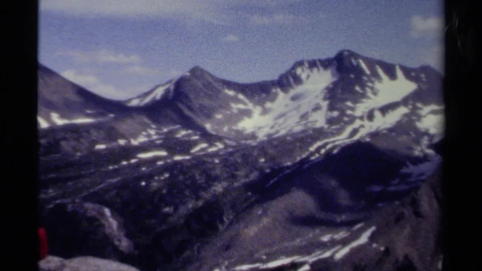 1979 8mm home-movie view of British Columbia alpine peaks with snow patches, rocky slopes, blue sky, and visible film gate edges.
