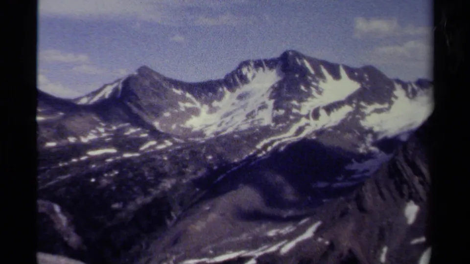 Vintage 8mm view of British Columbia mountains, showing snow-streaked peaks and dark alpine slopes beneath a clear blue sky.
