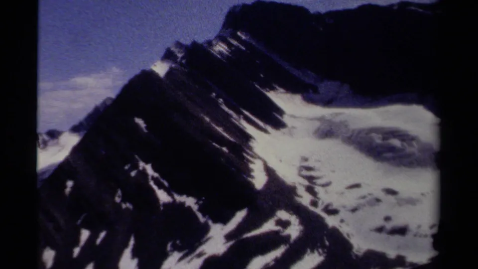 Close 8mm view of a steep British Columbia mountain face, with dark rock, bright snowfields, and visible home-movie grain.