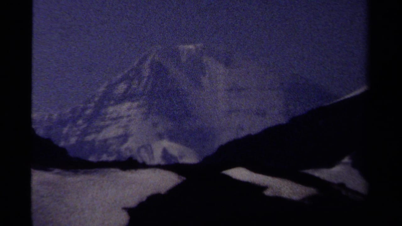 1979 British Columbia 8mm frame showing a snow-covered mountain peak behind dark ridges under a hazy blue sky.