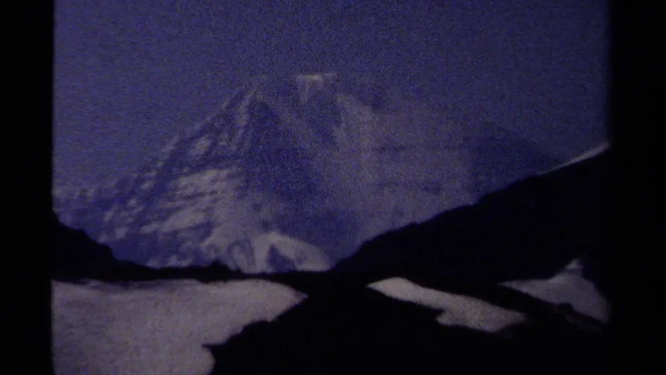 1979 British Columbia 8mm frame showing a snow-covered mountain peak behind dark ridges under a hazy blue sky.
