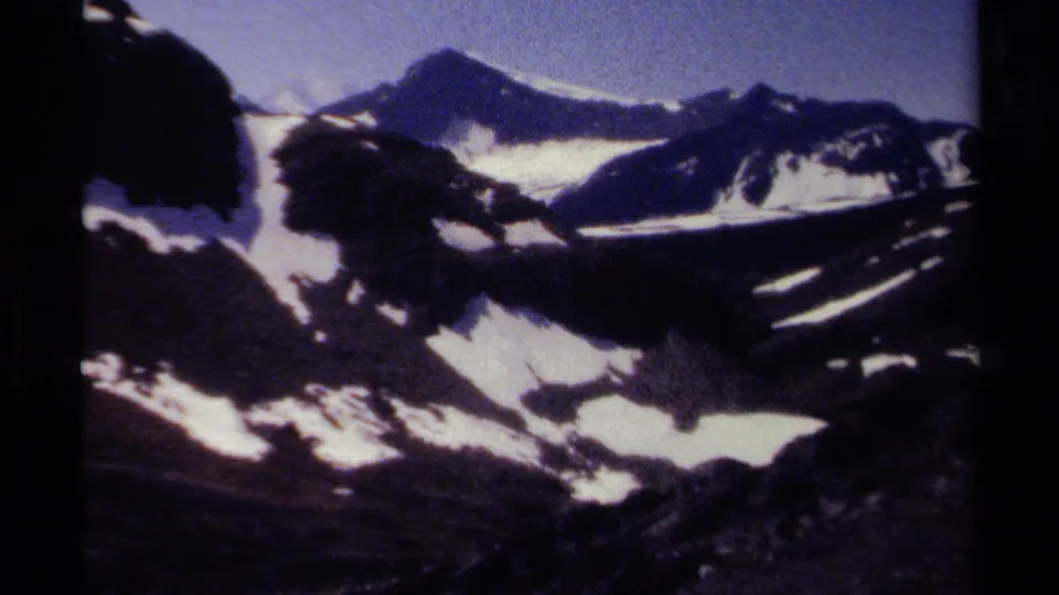 Wide 1979 British Columbia 8mm frame with snow bands across rugged dark mountains and a pale blue sky above.