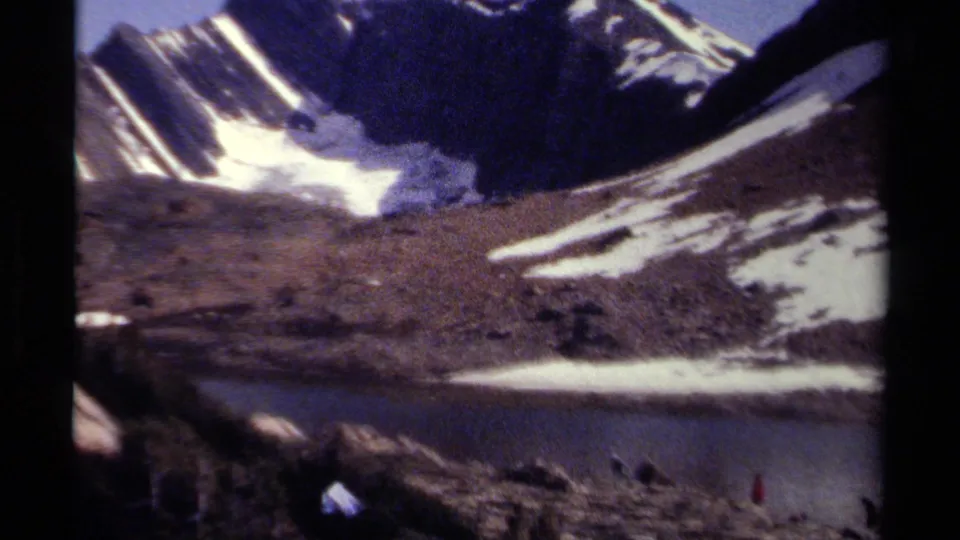 1979 British Columbia lakeside mountain view, with small hikers near the water below snow-streaked slopes and rocky shoreline.