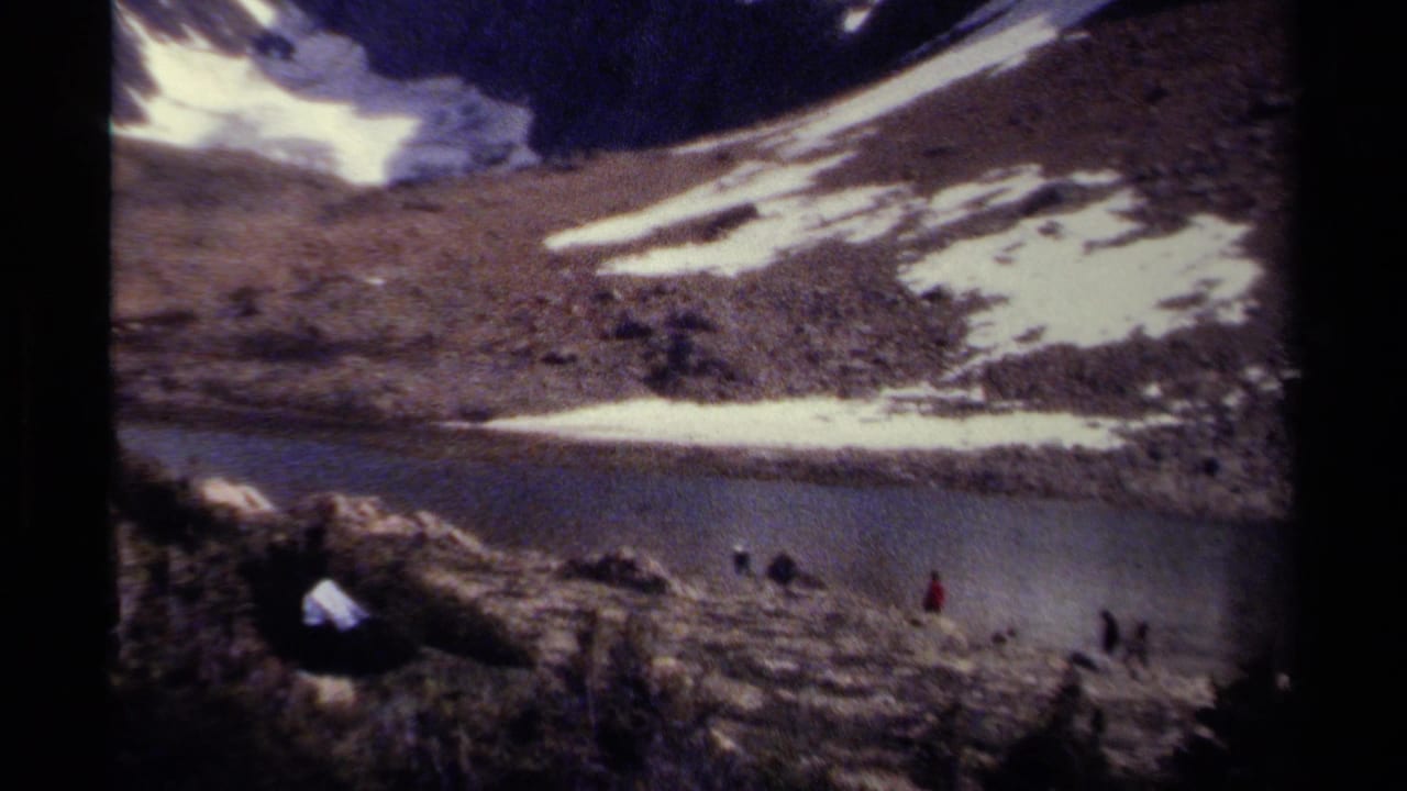 1979 British Columbia 8mm frame showing distant hikers crossing rocky terrain near a calm alpine lake and snow-streaked slope.