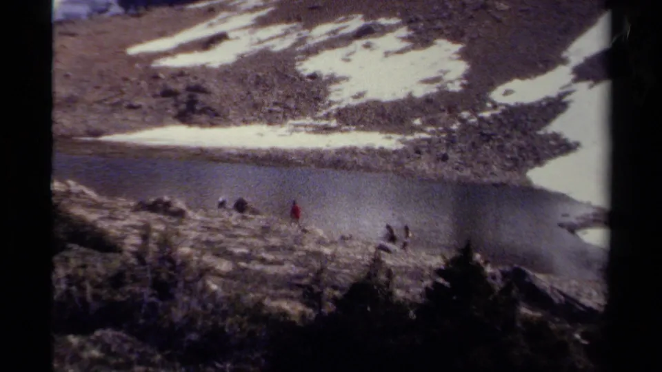 British Columbia home-movie frame of hikers moving across rough ground beside a still alpine lake and patchy snow.