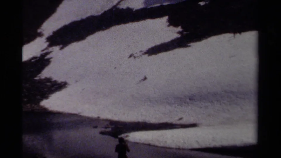 A distant figure stands near the bottom of an 8mm frame dominated by snowfields, dark rock, and alpine water.