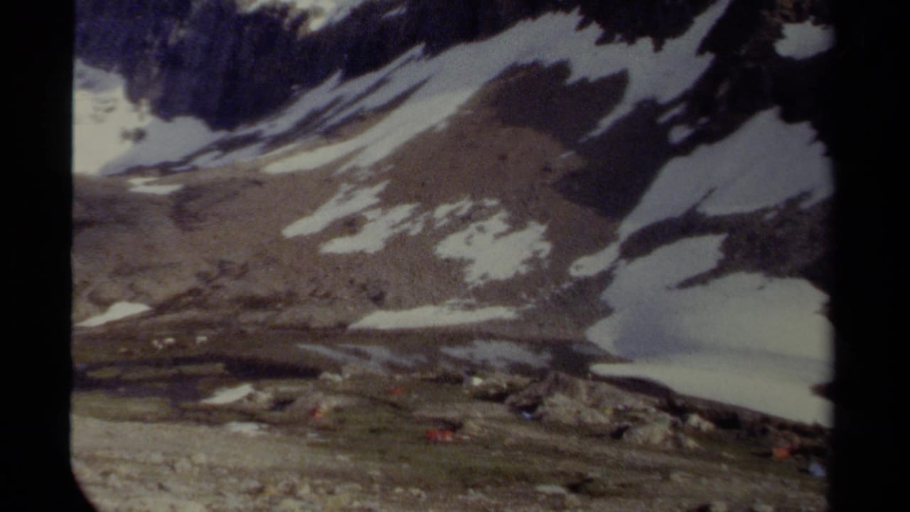 1979 8mm home-movie frame from British Columbia showing snow patches on a rugged mountain slope above rocky alpine terrain and a small lake area.