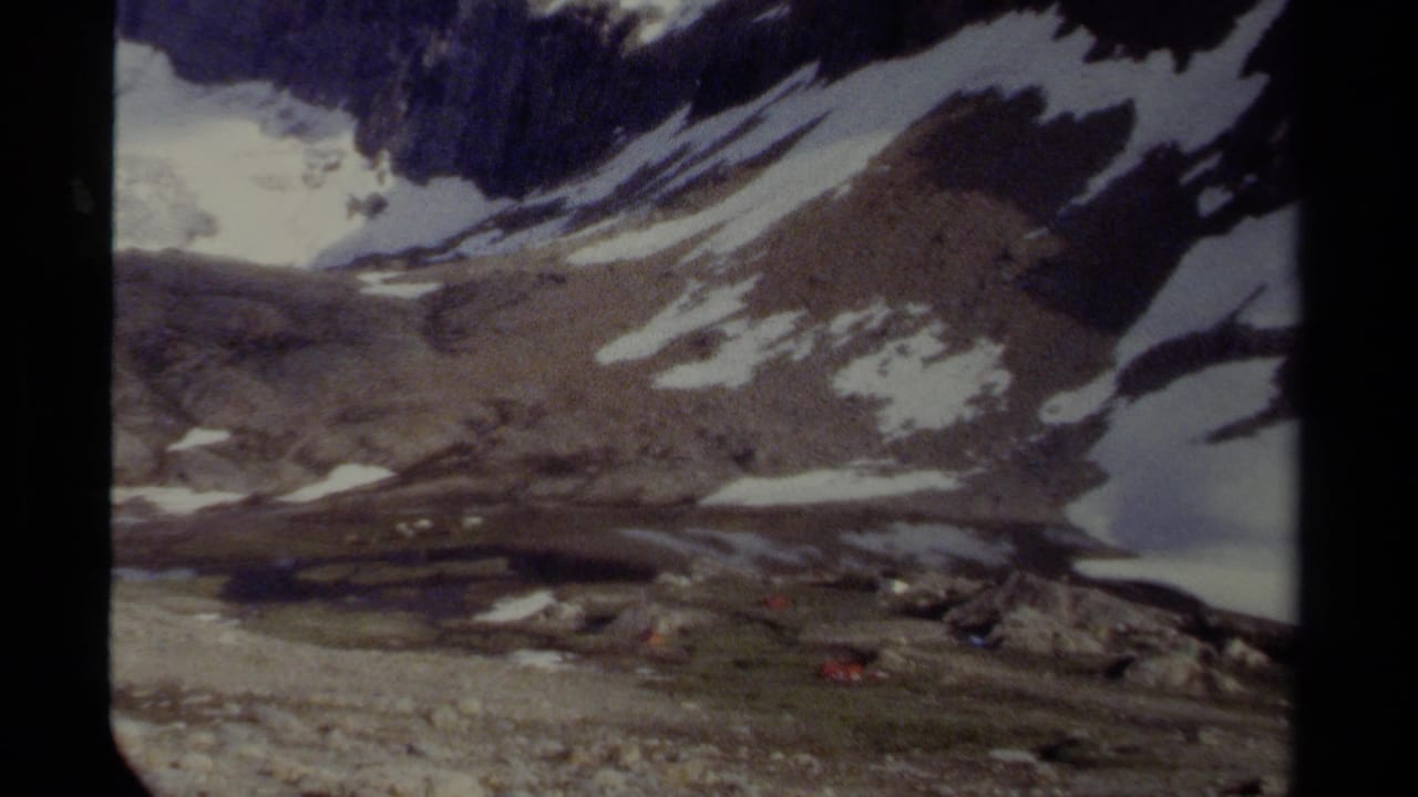 British Columbia alpine landscape on 8mm film, with snow-streaked slopes, rocky terrain and a small lake or meltwater basin near the mountain base.