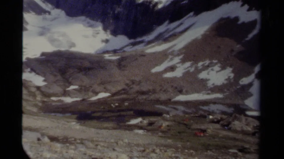 Vintage 8mm frame of a British Columbia mountain landscape, showing a broad rocky slope with scattered snow patches and sparse alpine vegetation.