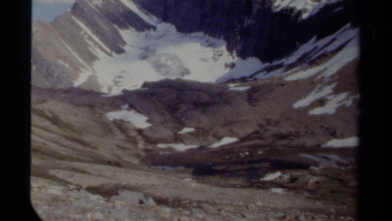 Snow-dusted mountain basin with rocky slopes, glacier ice, blue sky, and black 8mm frame borders.