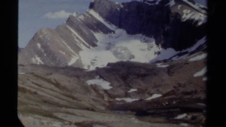 Vintage 8mm view of British Columbia alpine terrain, with diagonal rock strata, snow patches, glacier ice, and soft faded color.