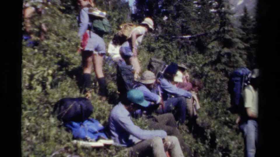 Group of friends rests beside a British Columbia mountain trail in 1979, with backpacks, hats, shrubs, and conifer trees visible.