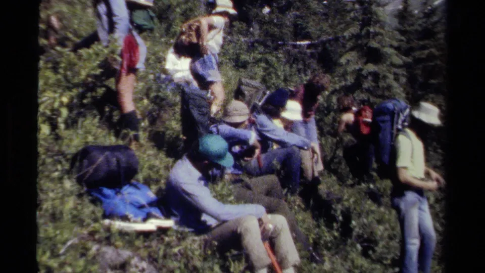 1979 8mm home-movie frame of hikers taking a break on a British Columbia trail, surrounded by brush, trees, and packs.