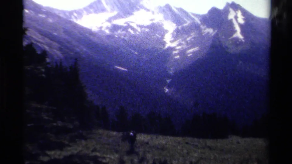 A handheld 8mm view in British Columbia shows snowy mountain ridges above dark evergreen forest and a small figure in the meadow below.