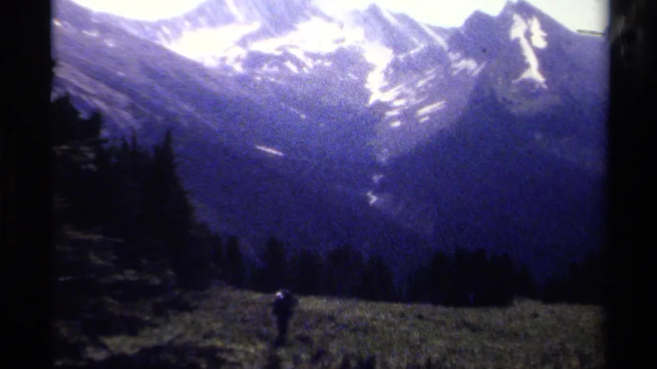 British Columbia mountain scenery fills the frame as a distant hiker appears in a meadow beneath snow-streaked ridges and dark forest.