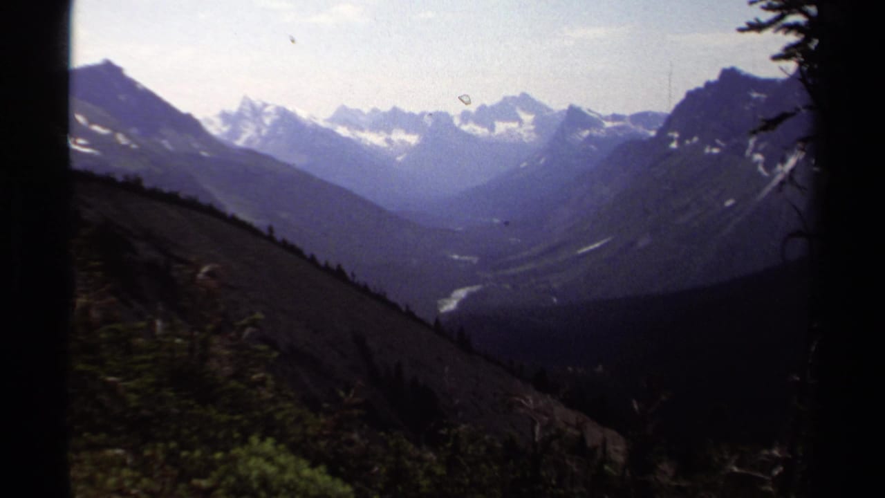 1979 8mm British Columbia view of snow-capped peaks, a forested valley, and a pale winding river seen from a high slope.