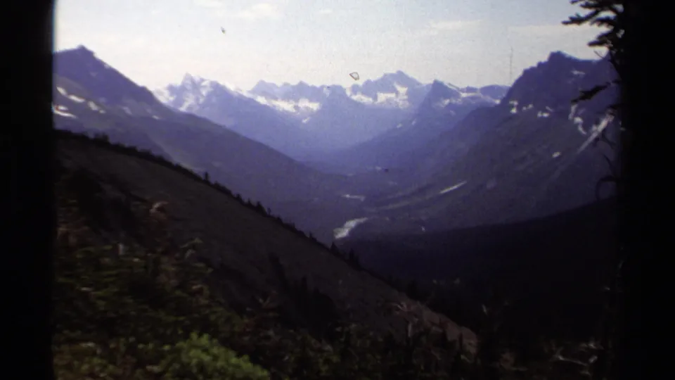 1979 8mm British Columbia view of snow-capped peaks, a forested valley, and a pale winding river seen from a high slope.